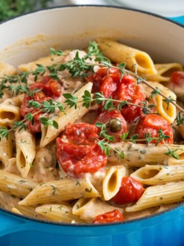 A close-up of creamy penne pasta in a blue pot, garnished with fresh thyme and cherry tomatoes. The rich sauce, possibly made with Boursin cheese, coats the pasta, creating a flavorful and comforting dish. This Boursin Cheese Pasta Recipe is a perfect combination of creamy texture and vibrant, fresh ingredients.