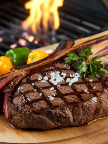 Grilled Tomahawk steak recipe featuring a perfectly seared steak with grill marks, topped with flaky sea salt, served alongside colorful roasted bell peppers, with a fire-lit grill in the background.