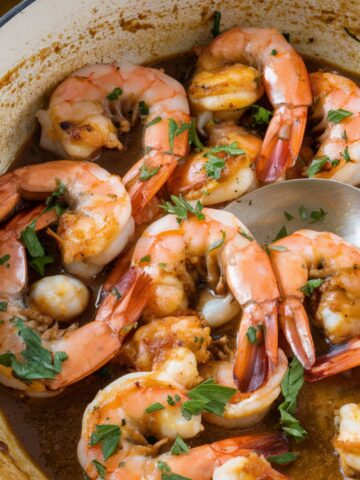 A close-up of a BBQ Shrimp Recipe, featuring plump shrimp cooked in a rich, buttery garlic sauce inside a blue enameled pan. The shrimp are coated in a flavorful BBQ seasoning and garnished with fresh parsley. The dish has a golden-brown sear, with a spoon lifting some shrimp, showcasing their juicy texture. Perfect for a Southern-style seafood meal.