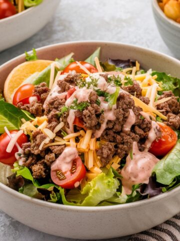 A loaded burger bowl recipe featuring seasoned ground beef on a bed of fresh lettuce, cherry tomatoes, shredded cheddar cheese, and a drizzle of creamy special sauce. Garnished with fresh herbs, this protein-packed, low-carb meal is a delicious and healthy alternative to a classic cheeseburger.