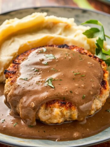 "Delicious Salisbury Steak Recipe With Ground Chicken, smothered in a rich and creamy mushroom gravy, garnished with fresh parsley, and served with roasted sweet potatoes and steamed broccoli on a white plate.