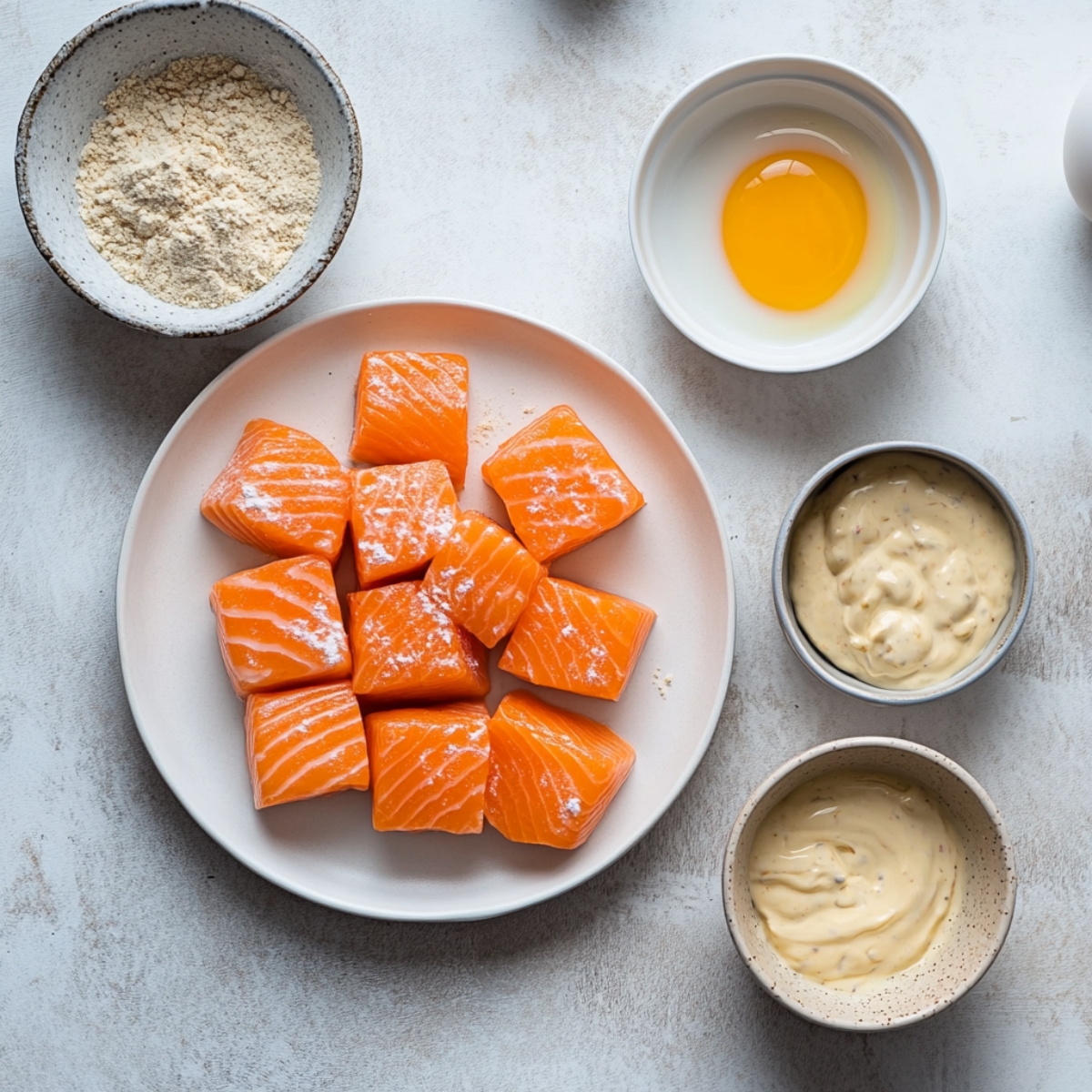 Top-down view of cubed raw salmon and bowls of flour, eggs, panko, and sauce ingredients on a white kitchen counter.