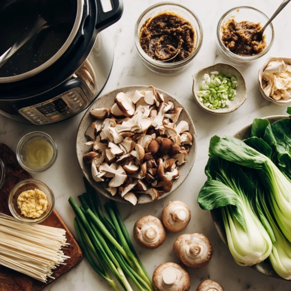 Overhead view of chopped vegetables, minced garlic, miso paste, and ginger on a white kitchen counter beside an open Instant Pot on sauté mode.