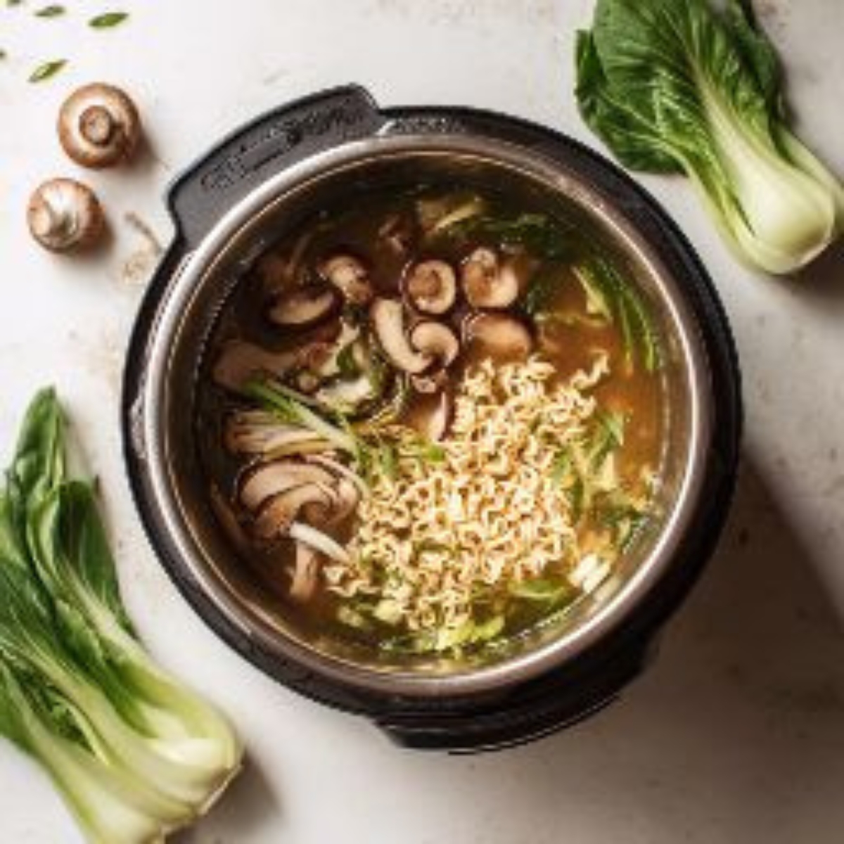 Closeup overhead view of mushrooms and dry ramen noodles placed in broth inside the Instant Pot, surrounded by chopped veggies on counter.