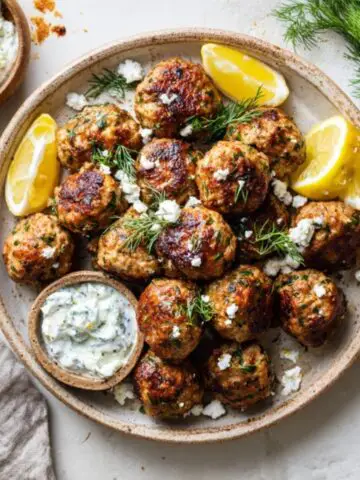 Plate of homemade Greek turkey meatballs with tzatziki sauce and lemon wedges on a white kitchen counter.