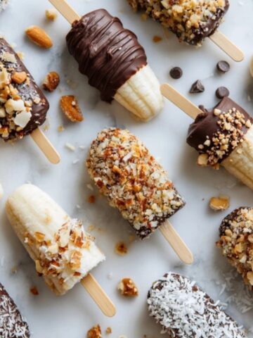 Homemade frozen banana snacks dipped in chocolate, nuts, and coconut on a white marble counter