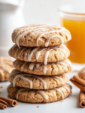 Soft glazed apple cider cookies stacked beside a glass of cider on a white kitchen counter.